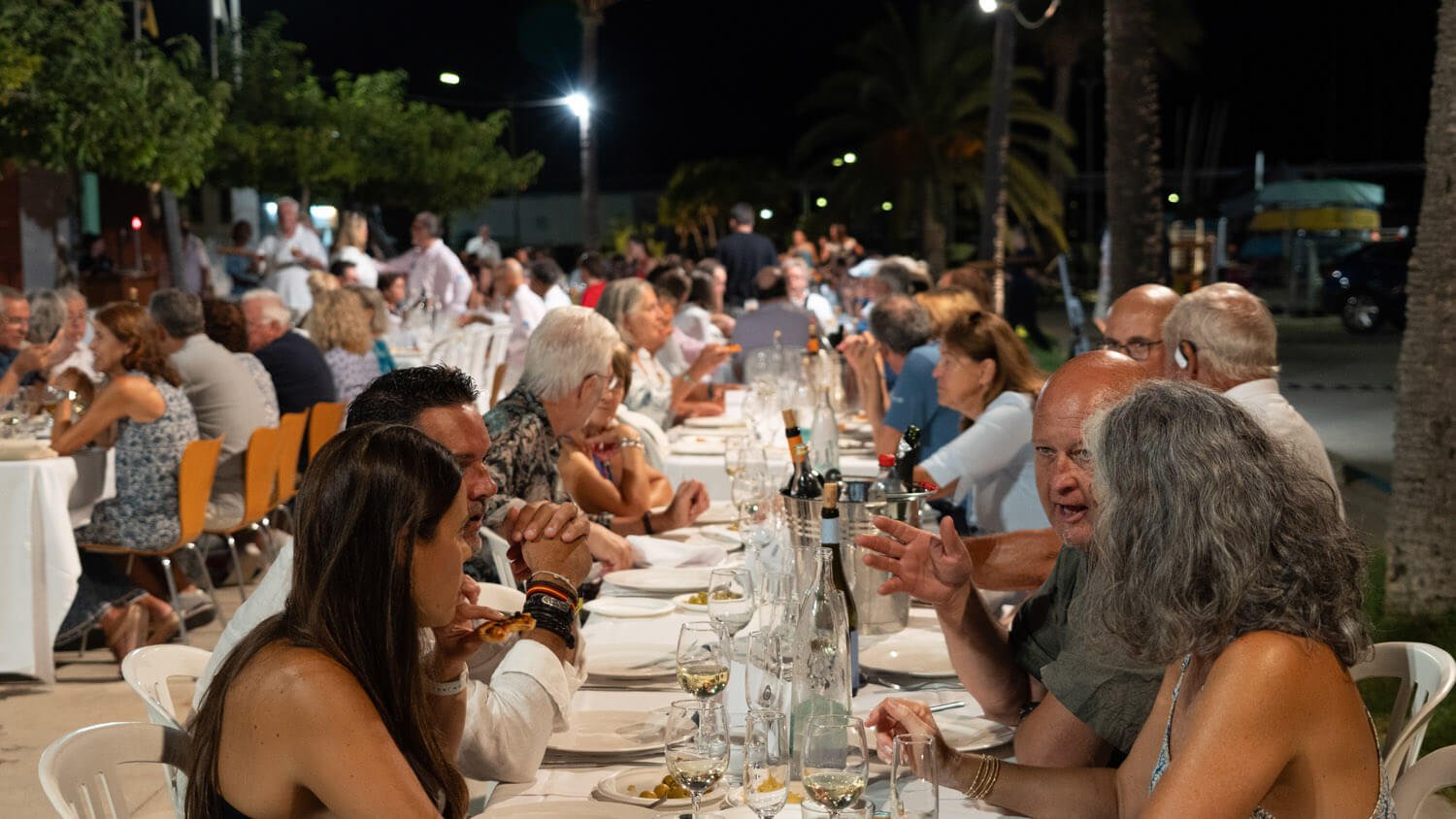 Grupo de personas cenando al aire libre en una mesa larga en la noche, con árboles y palmeras en el fondo.