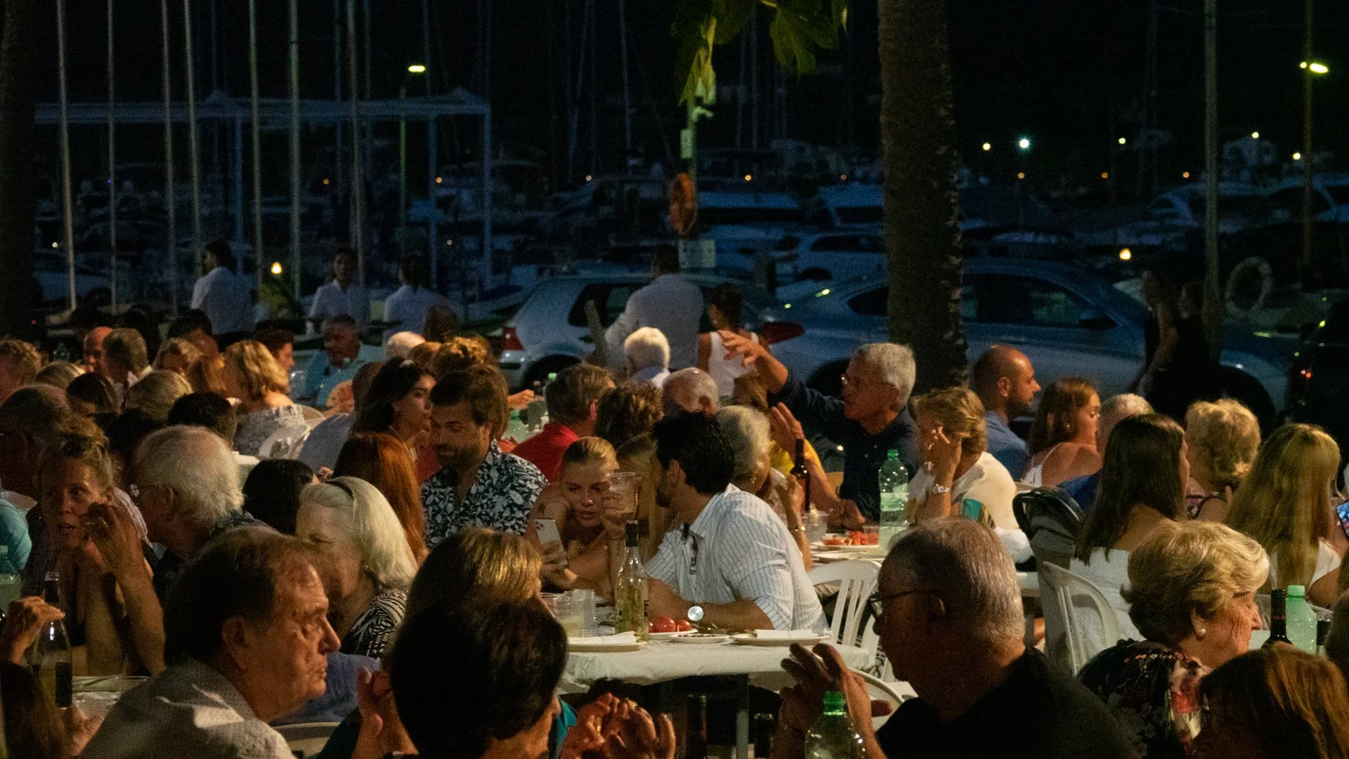 Reunión o fiesta al aire libre por la noche con muchas personas sentadas en mesas, conversando y disfrutando, en un entorno con árboles y muchas embarcaciones en un puerto en el fondo.