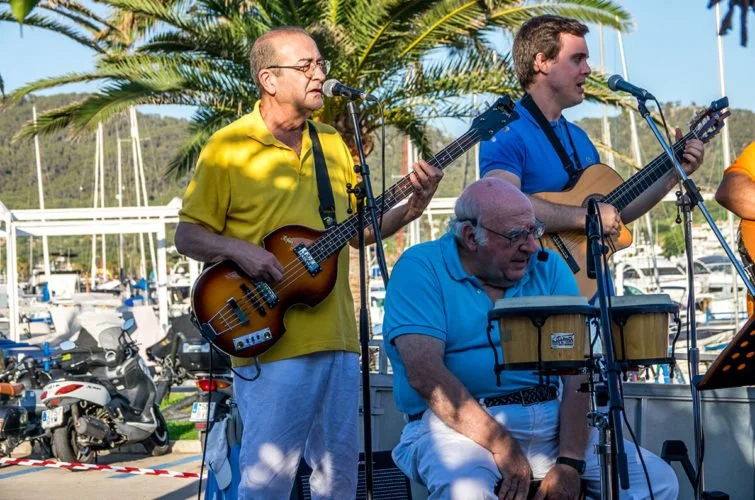Tres músicos tocando en un escenario al aire libre, con yates y palmeras de fondo, en un día soleado.