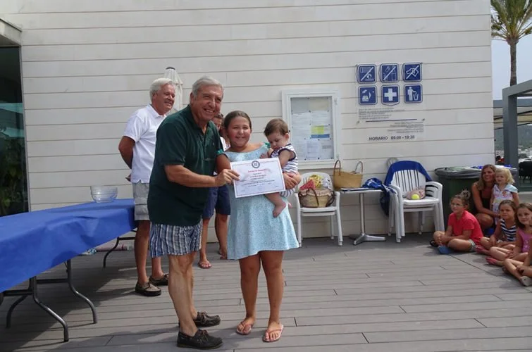 Una niña recibe un certificado de reconocimiento de un hombre mayor en un evento al aire libre con varias personas sentadas en el suelo y un grupo de niños en el fondo.