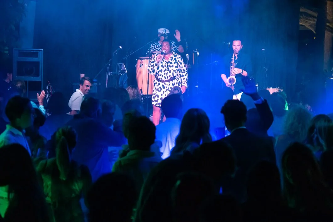 Un grupo de personas disfrutando una presentación en un escenario con música en vivo, con una cantante y músicos tocando el saxofón y la percusión, todo en un ambiente oscuro con iluminación de colores.