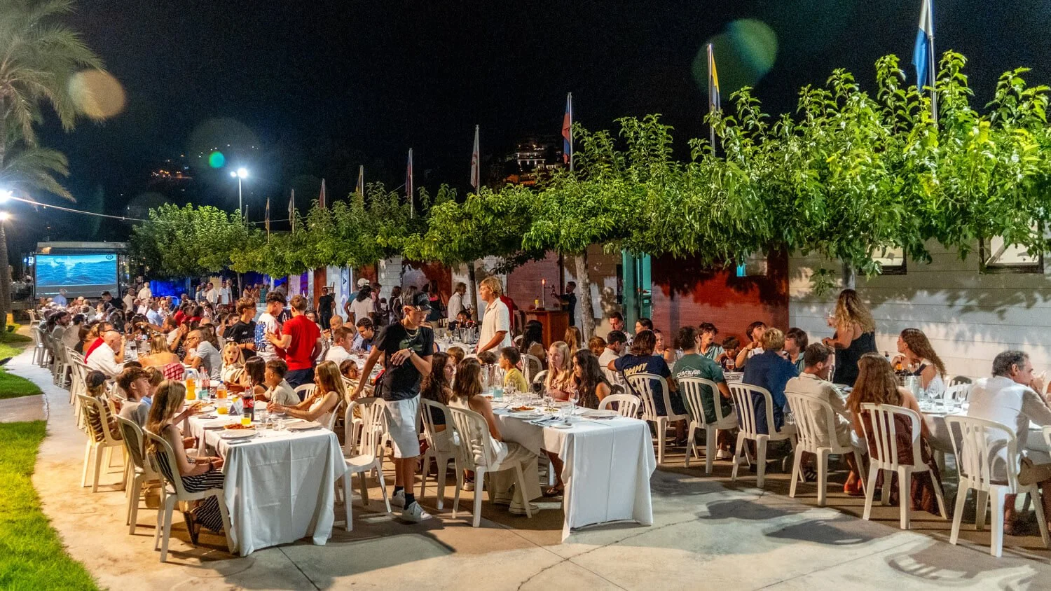 Una cena al aire libre en la noche con muchas mesas y personas disfrutando de la comida en un espacio festivo, decorado con árboles y banderas, en un ambiente alegre y concurrido.