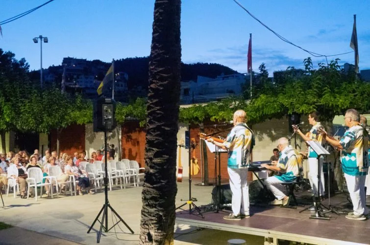 Conjunto musical tocando en un escenario al aire libre en la noche, con público sentado y viendo el espectáculo.