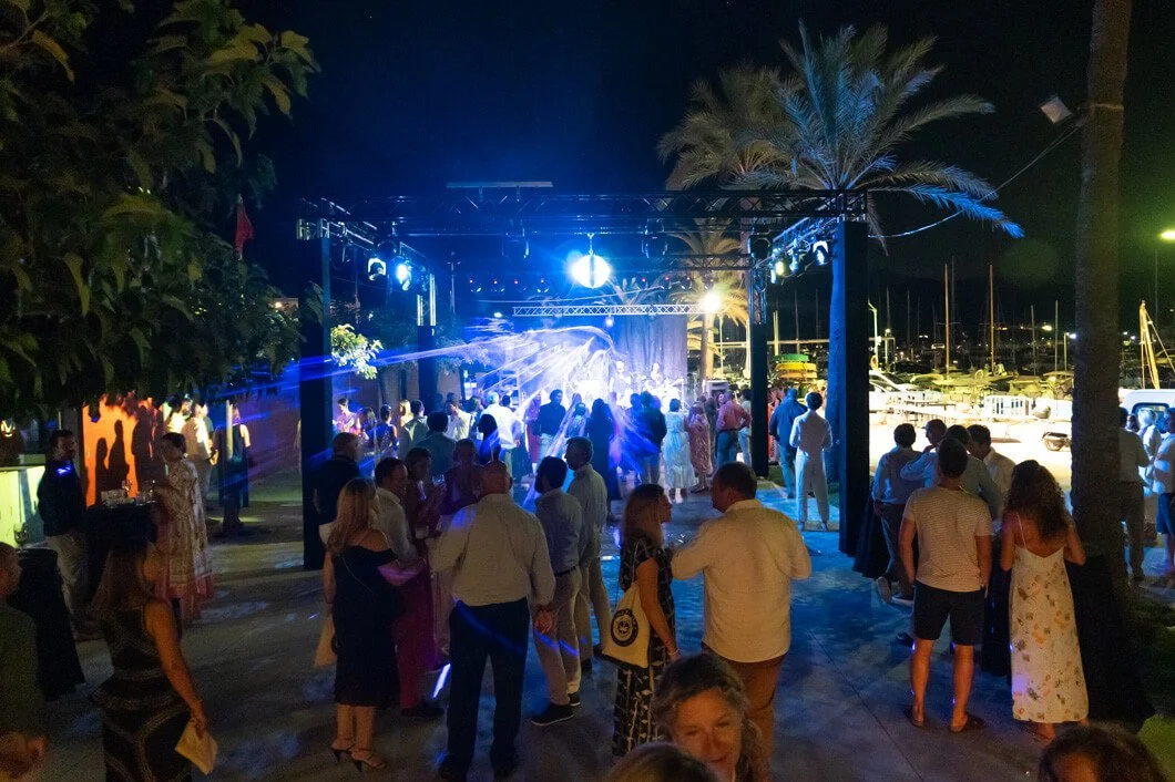 Personas bailando en una fiesta al aire libre con música y luces, en un entorno moderno con árboles y yates en el fondo.
