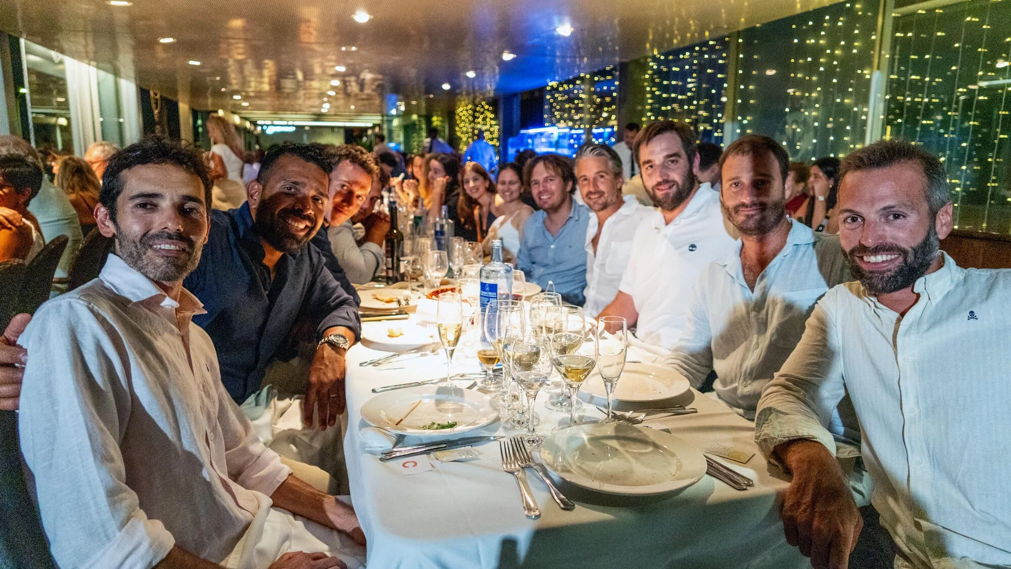 Grupo de personas sonriendo durante una comida en un restaurante decorado con luces. Hay varias botellas y copas de vino en la mesa.