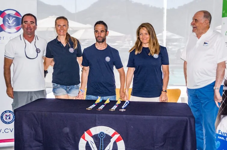 Cinco personas de pie detrás de una mesa cubierta con una bandera con logotipo de un ancla y remos. Tres de ellos llevan camisetas azul marino con logotipo, dos usan ropa blanca. En la mesa hay cuatro chelas y una botella.