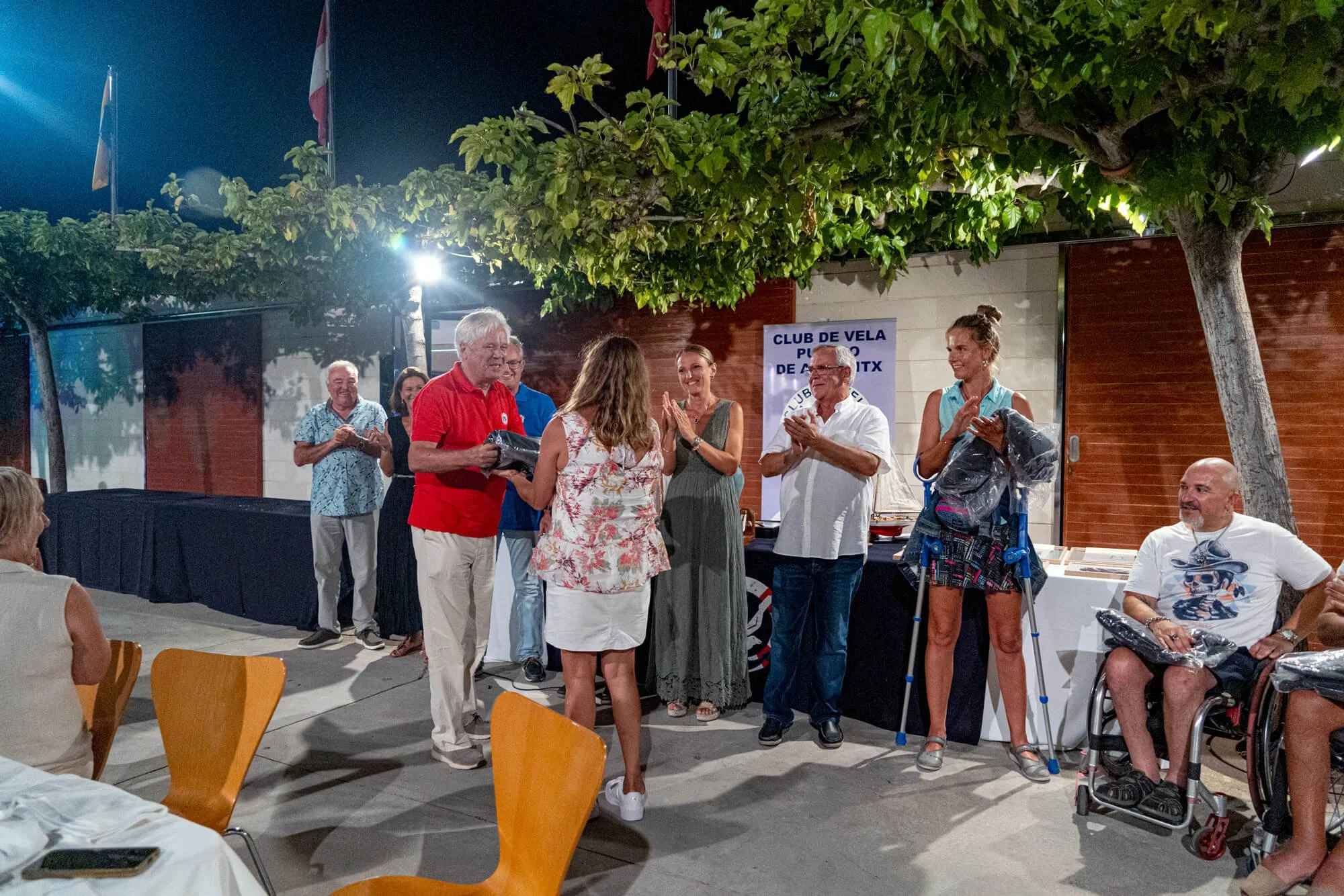 Grupo de personas en un evento al aire libre en la noche, algunas entregando premios, otras aplaudiendo y disfrutando de la ocasión, con un cartel de fondo que dice 'Club de Vela Puerto de Mazatlán'.