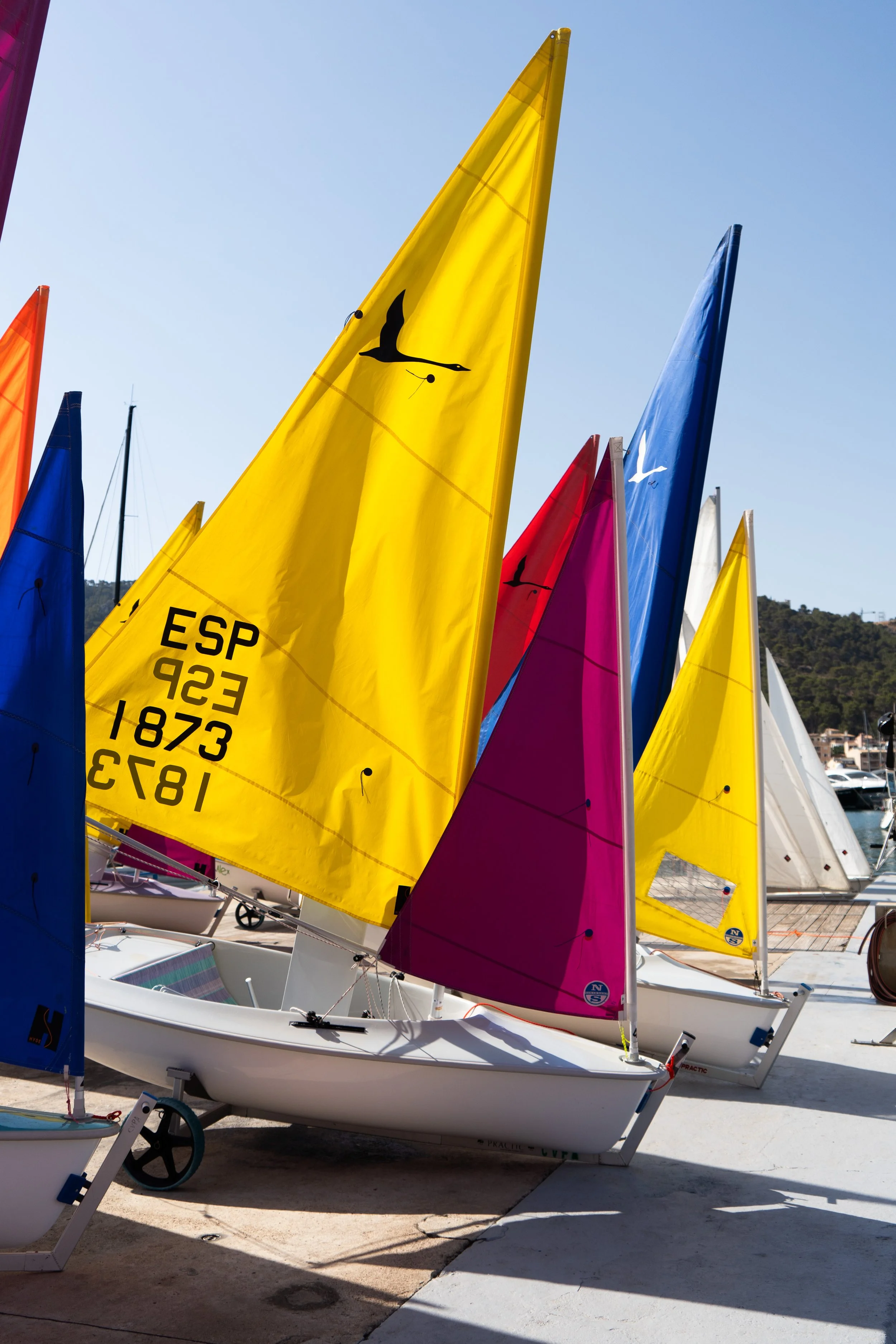 Vela de vela blanca en un bote en un muelle, con varias velas de diferentes colores (amarilla, rosa, azul, blanca) en primer plano. Fondo de cielo despejado y colinas.