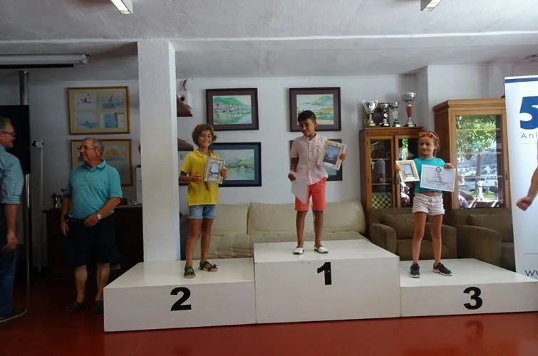 Niños en un podio de ganador, en una ceremonia de premiación, con niños en las posiciones 1, 2 y 3, sosteniendo diplomas, en un ambiente interior con cuadros en la pared y una vitrina con trofeos.