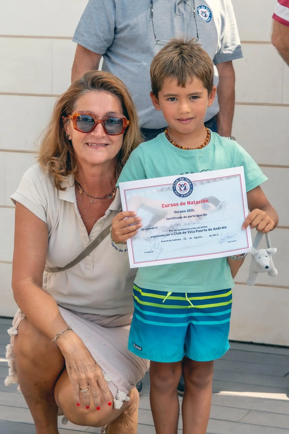 Niña sosteniendo un certificado de participación en cursos de natación, acompañado de una mujer con gafas de sol y otra persona parcialmente visible en el fondo.