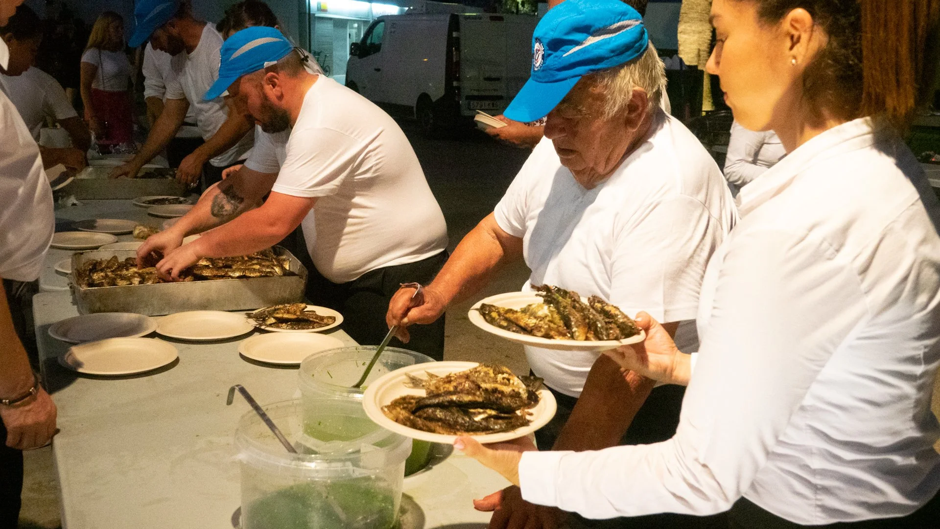 Personas sirviendo y comiendo pescado en una mesa en una especie de evento o comedor comunitario, en un ambiente nocturno.