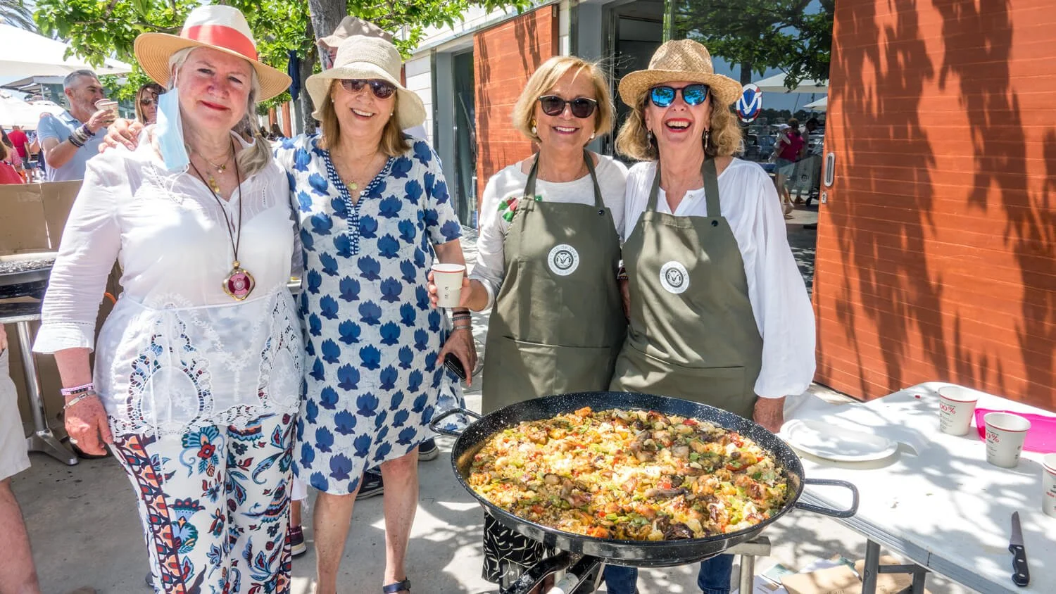 Cuatro mujeres sonrientes en un evento al aire libre, con un plato grande de paella en el centro, dos de ellas usan delantales y sombreros de paja, otras dos usan blusas diferentes, en un fondo de árboles, edificios y otras personas disfrutando.
