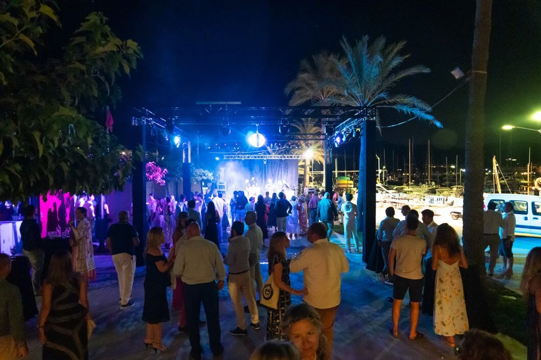 Fiesta al aire libre de noche con gente bailando y un escenario iluminado, cerca de barcos en un puerto.
