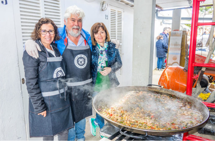 Tres personas sonrientes, dos mujeres y un hombre, cocinando en una plancha gigante con ingredientes como carne y verduras, en un evento al aire libre.