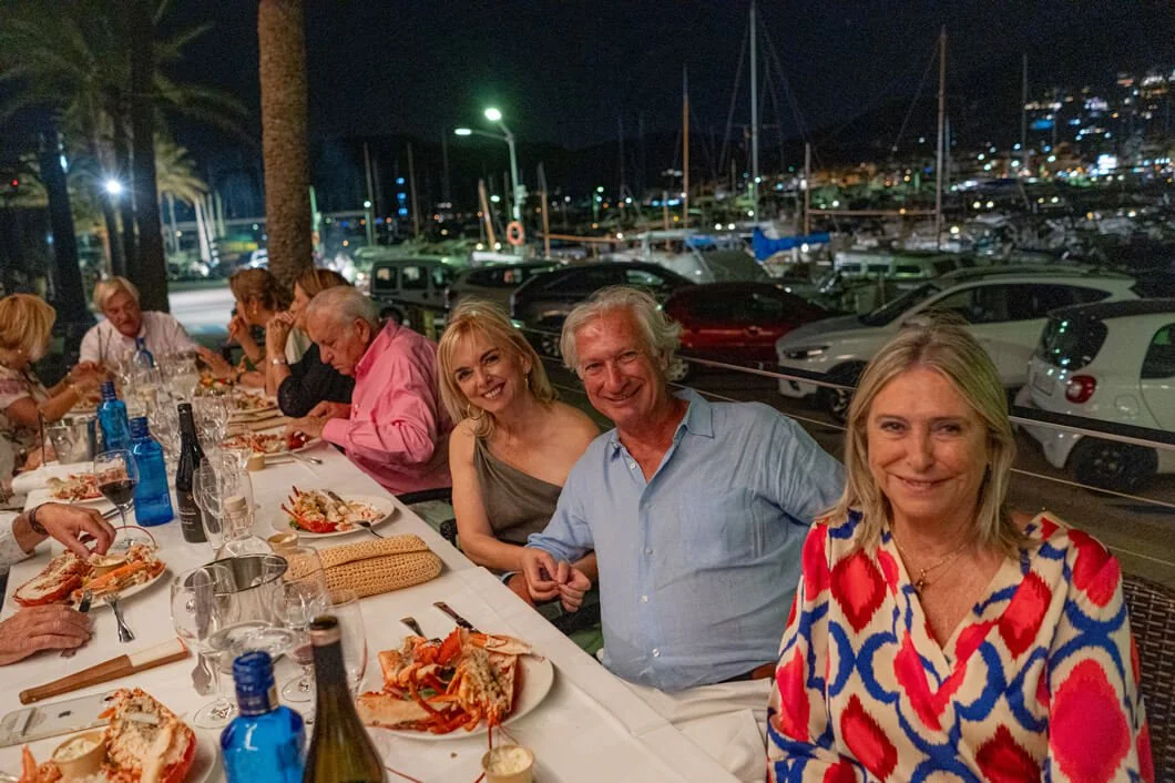Grupo de personas disfrutando de una cena al aire libre en un muelle, con barcos y autos al fondo, durante la noche.