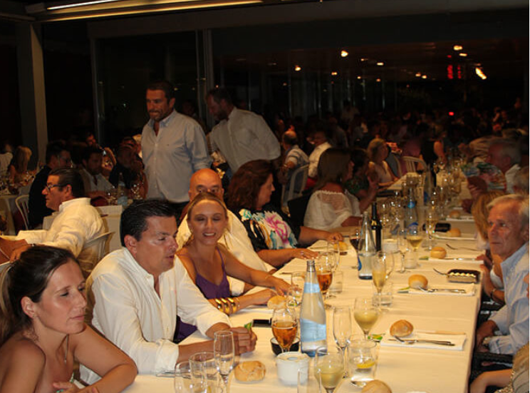 Personas sentadas en una mesa en un restaurante durante una comida, con comida y bebidas en la mesa, ambiente interior concurrido y bien iluminado