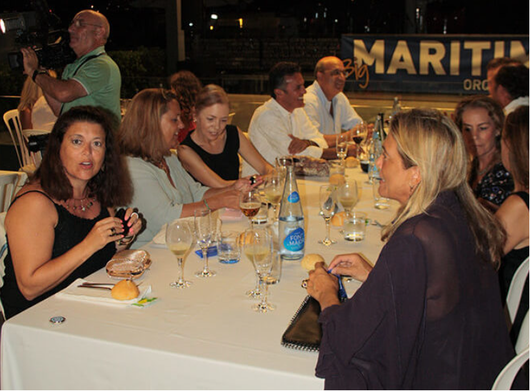 Grupo de personas sentadas en una mesa en un evento nocturno, algunas tomando fotos y otras conversando, con un cartel que dice 'Maritim'.