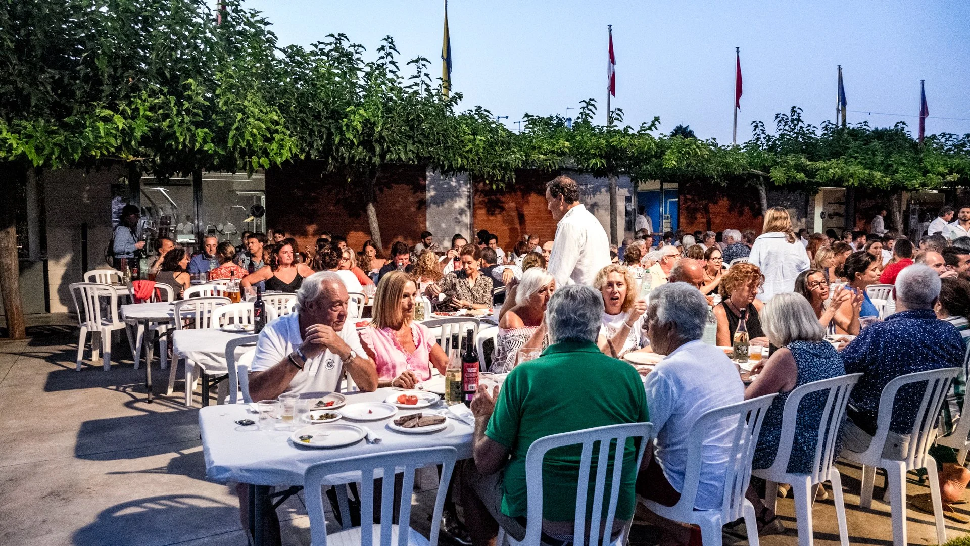Una multitud de personas cenando en una celebración al aire libre, con árboles y banderas en el fondo, en horario nocturno.