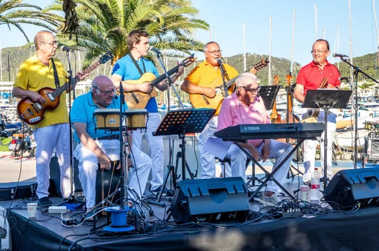 Grupo de músicos tocando instrumentos en un escenario al aire libre con fondo de palmeras y barcos.