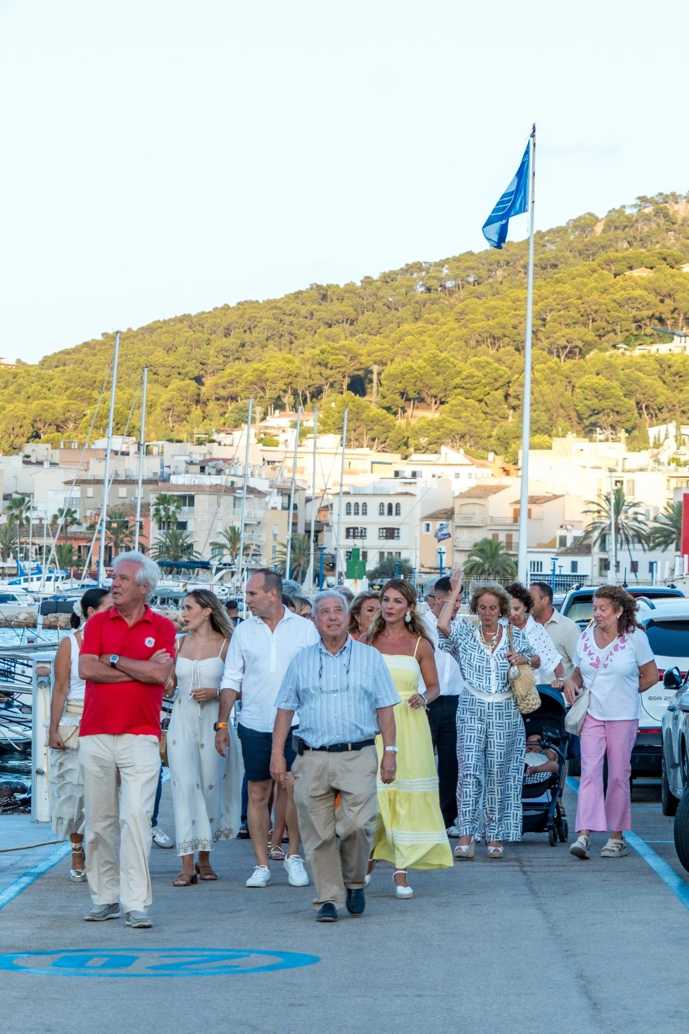 Grupo de personas caminando por un muelle cerca del mar, con casas y colinas verdes al fondo.