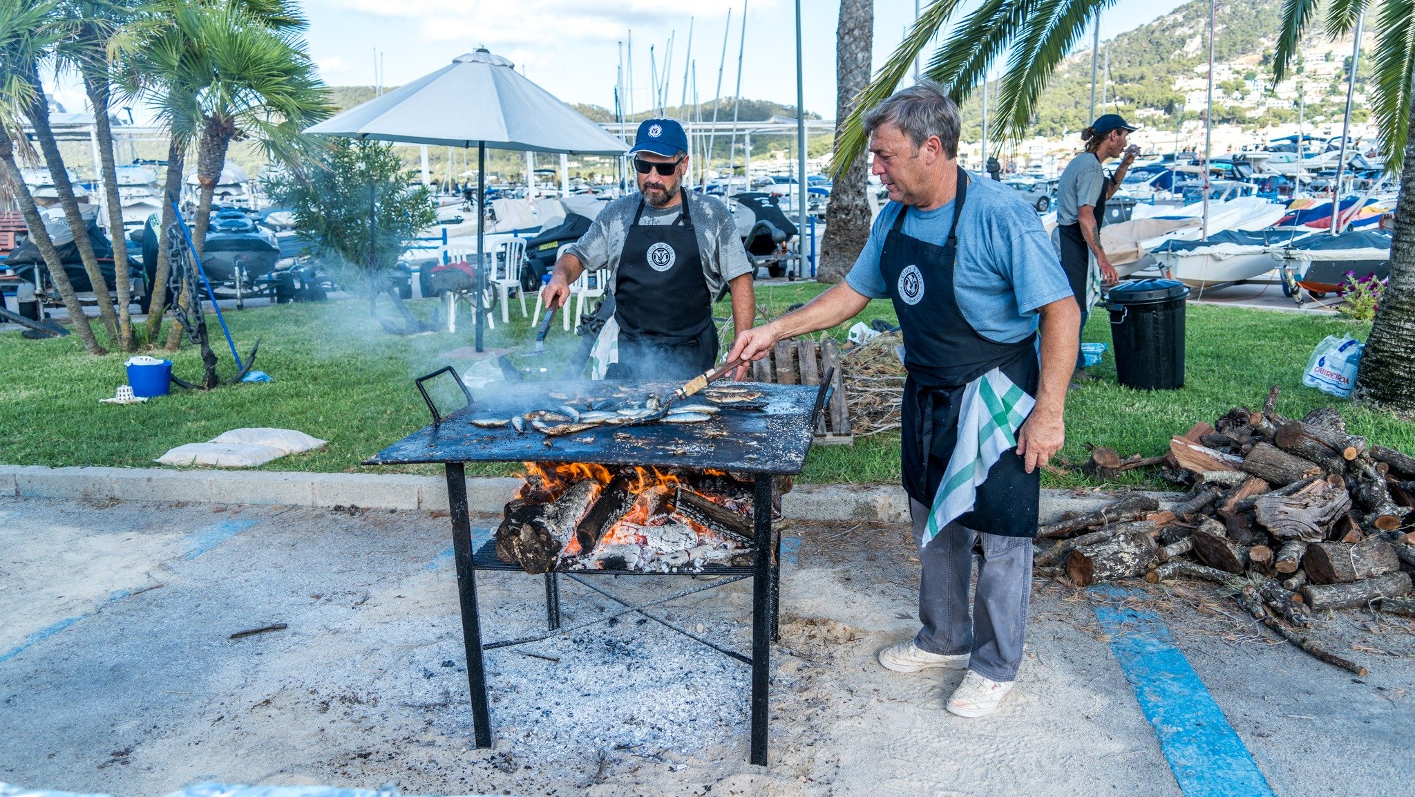 Dos hombres cocinando mariscos en una parrilla al aire libre cerca de un puerto con yates y palmeras.