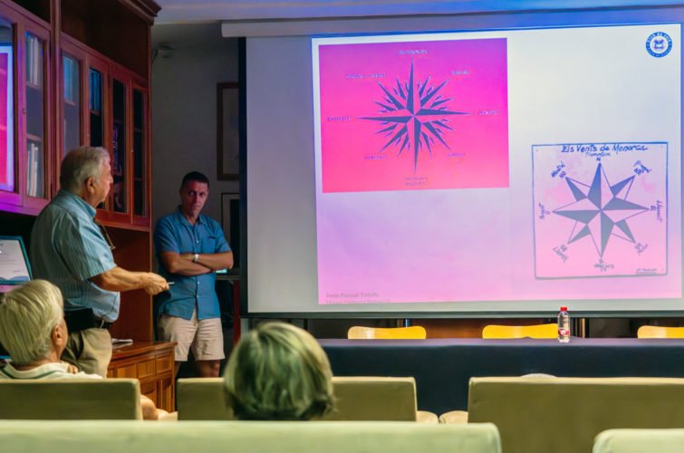 Varias personas en una sala de reuniones viendo una presentación sobre una rosa de los vientos en una pantalla grande.