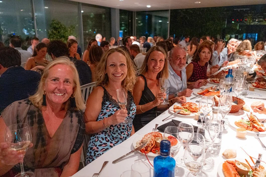 Grupo de personas en una cena elegante, sonriendo y brindando con copas de vino, en un ambiente social y festivo.