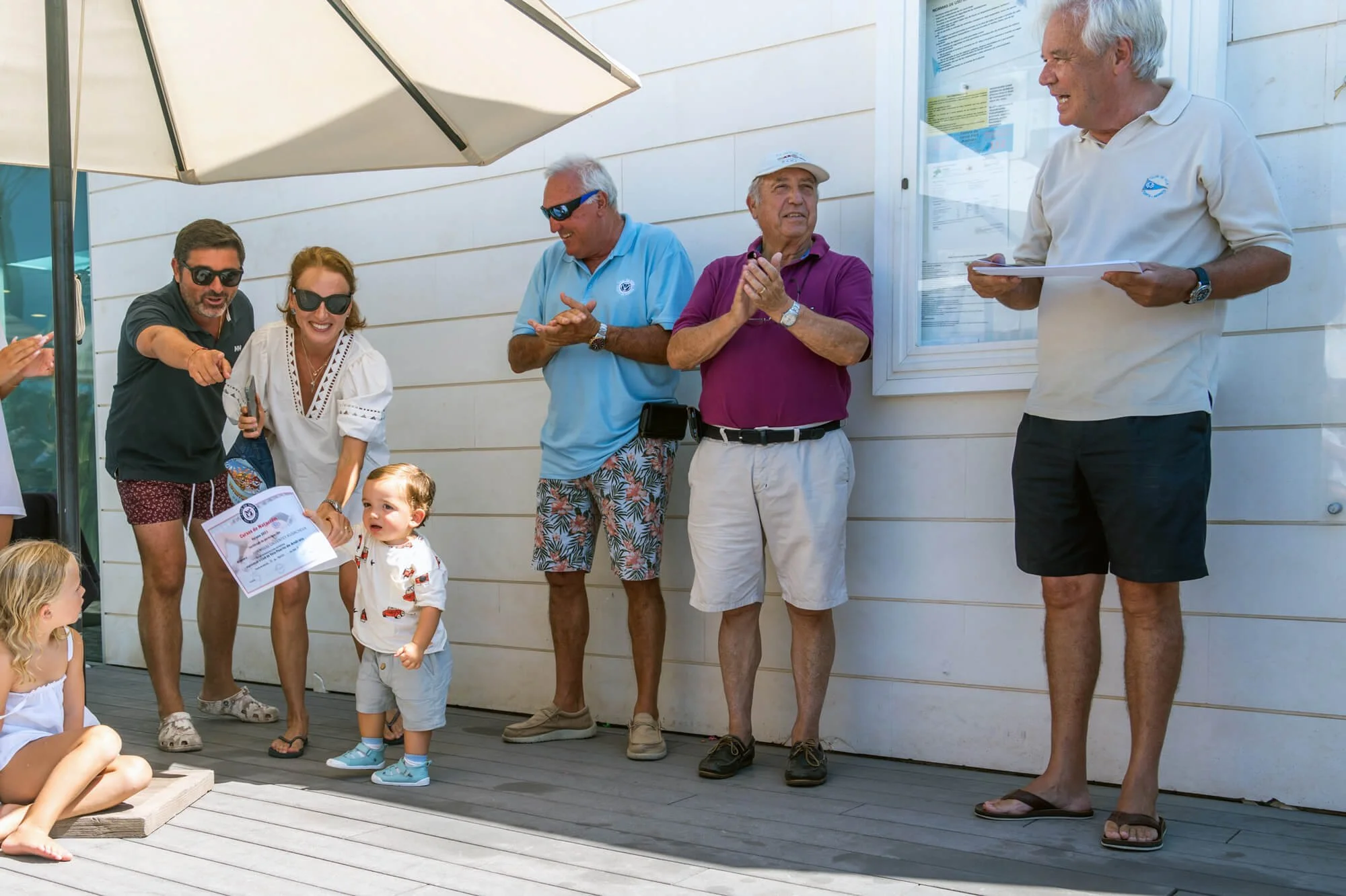 Grupo de personas en un evento al aire libre, algunos niños sentados y adultos de pie, dos de los adultos llevan gafas de sol, hay un hombre con camisa blanca y un documento en las manos, otros adultos parecen estar recibiendo un reconocimiento o cer