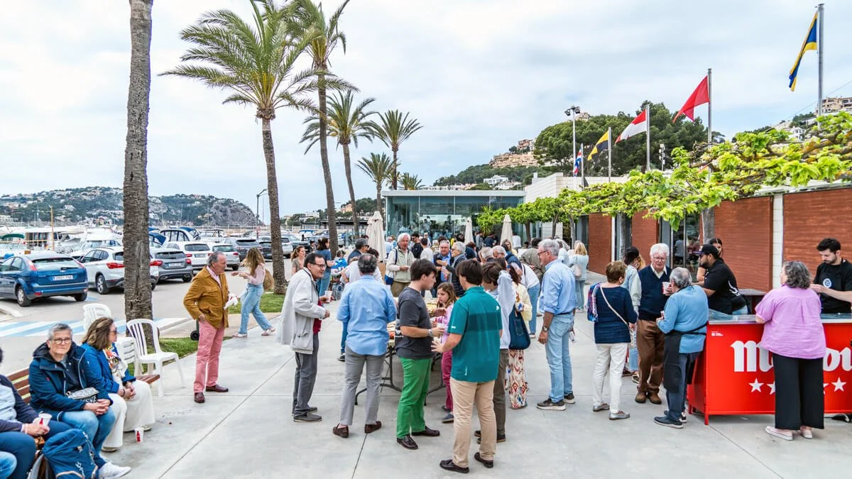 Gente en fila disfrutando de un evento al aire libre en un muelle con palmeras, cerca del mar, con banderas y un toldo rojo que dice "mundo".