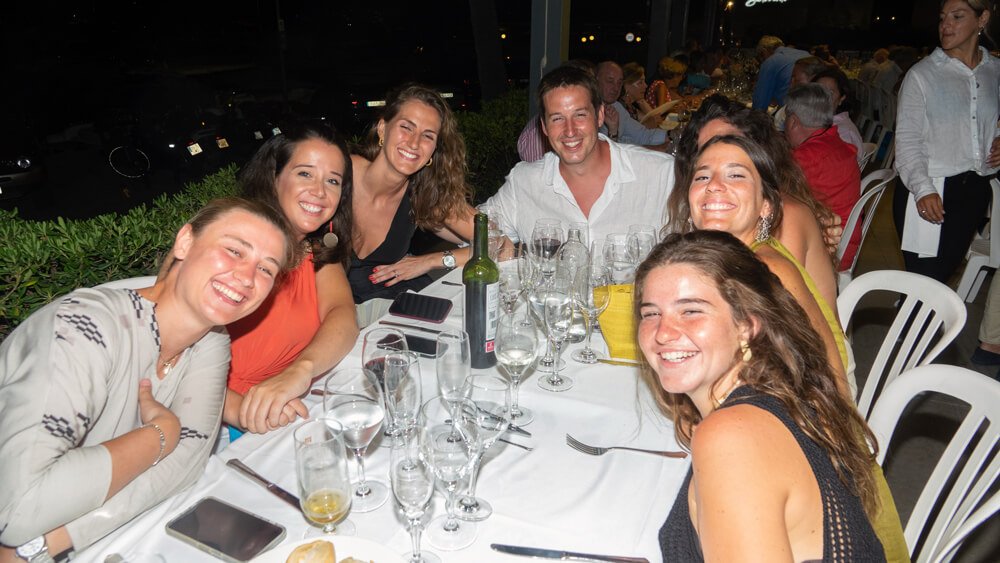 Grupo de seis personas sonriendo en una cena al aire libre con muchas copas de vino y comida en una mesa