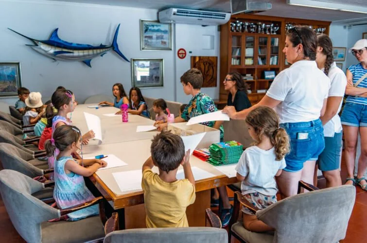 Niños y adultos en una sala participando en una actividad o clase, alrededor de una mesa grande con papeles y libros, con decoración marina en la pared.
