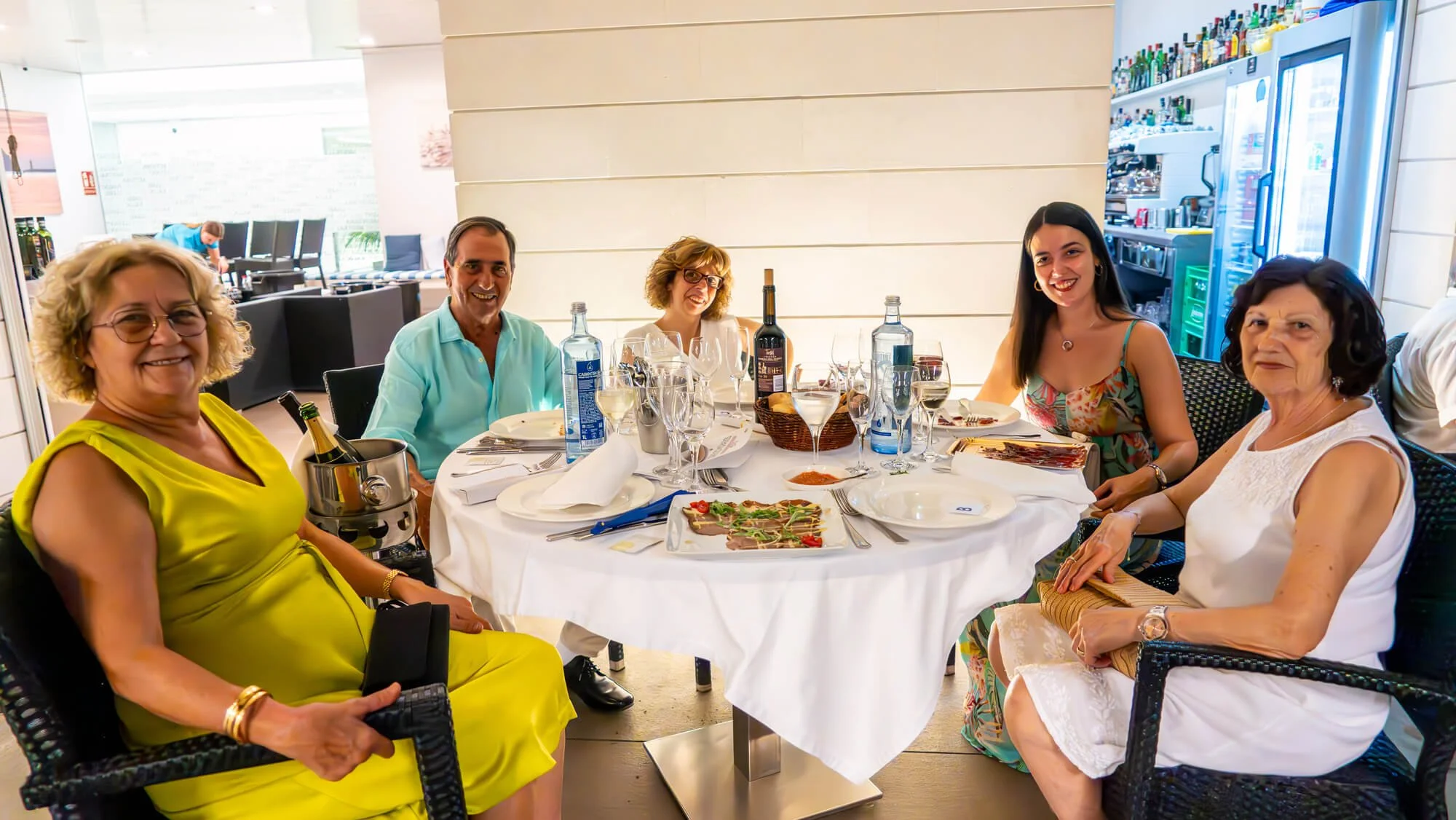Grupo de cinco personas sentadas en una mesa de restaurante disfrutando de comida y bebida, sonriendo y mirando hacia la cámara.