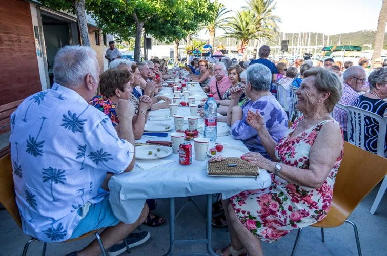 Un grupo de adultos mayores sentados en una mesa larga al aire libre, disfrutando de una comida en un entorno con árboles y palmeras.