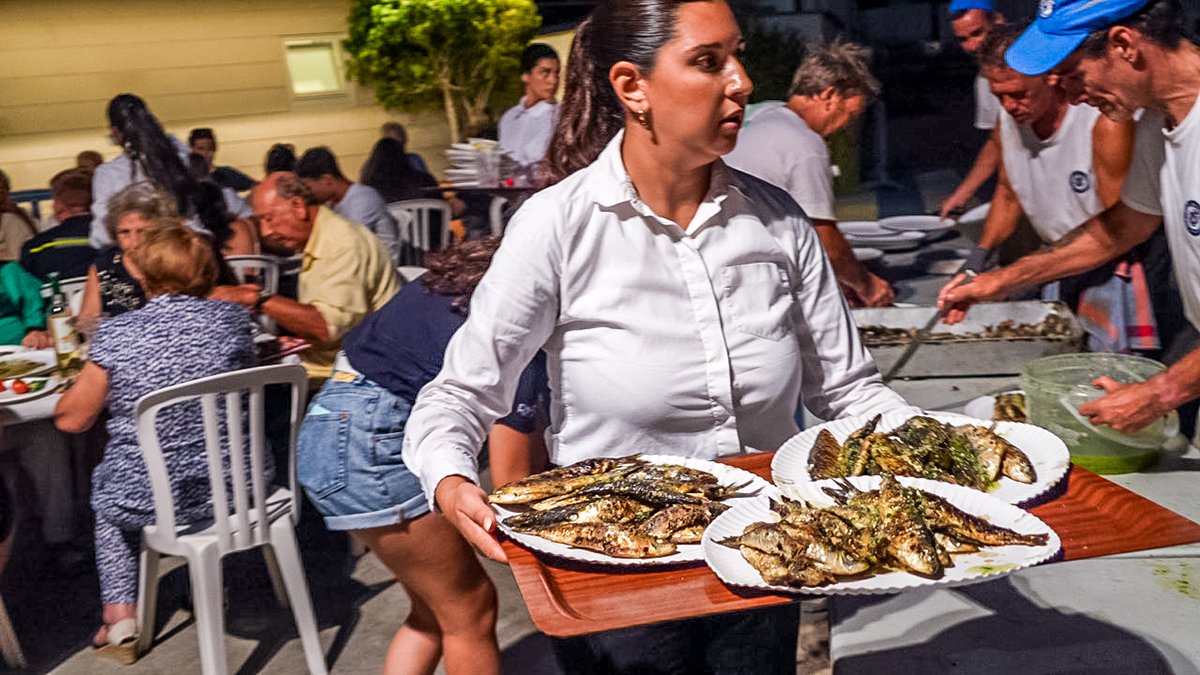 Una mujer sostiene una bandeja con platos de pescado asado en una reunión al aire libre, con muchas personas sirviendo comida en el fondo.
