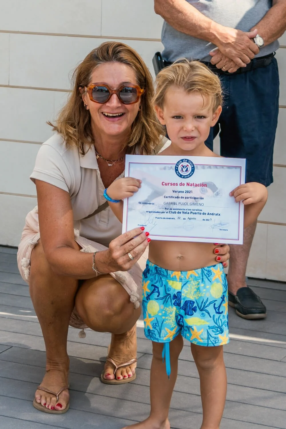 Niño con certificado de participación en cursos de natación, acompañado por una mujer sonriente con gafas de sol y cabello rizado, en un evento de verano en Puerto de Andratx.