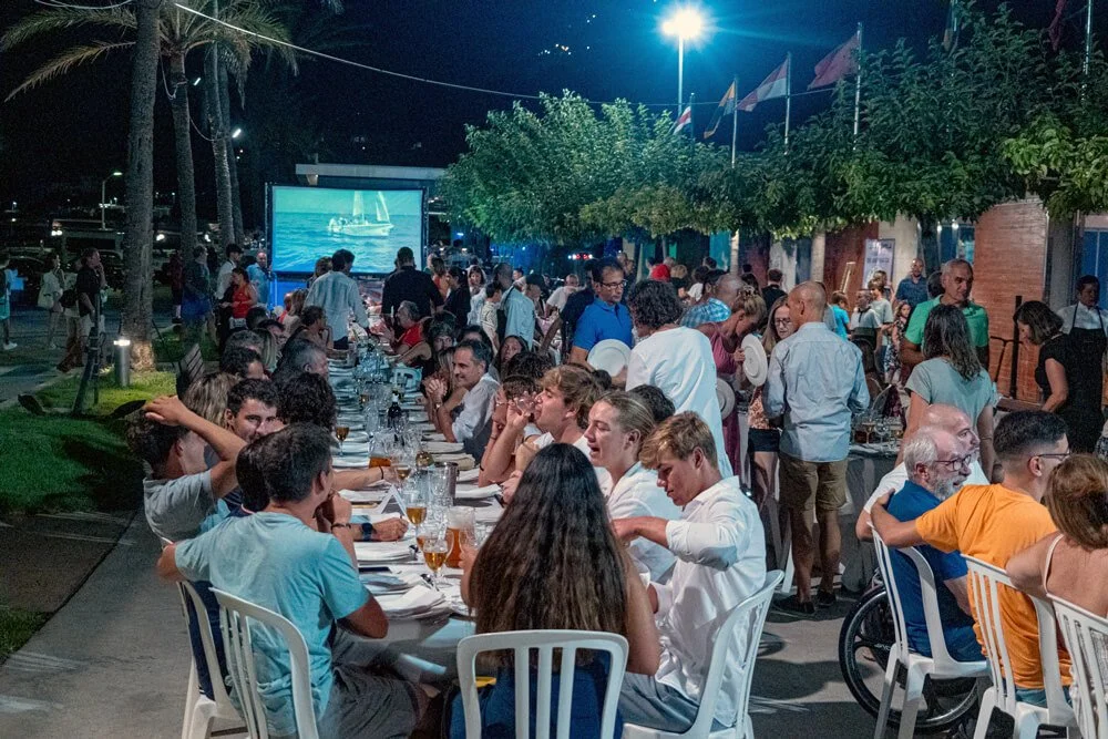 Una reunión social en exteriores con muchas personas sentadas en una mesa larga, disfrutando de comida y bebida, mientras otras se reúnen de pie y observan una pantalla gigante que muestra un partido de fútbol, con árboles y banderas en el fondo de n