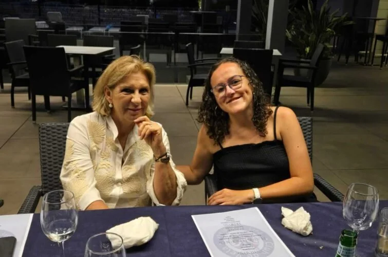 Dos mujeres sentadas en una mesa de restaurante, una joven con camiseta negra y gafas, y una mujer mayor con blusa blanca y cabello rubio. La mesa tiene copas de agua y papel.