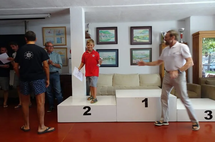 Premiación en un evento con niños en un podio, niño en tercer lugar con una medalla, un adulto en primer lugar haciendo una señal y otros niños y adultos observando.