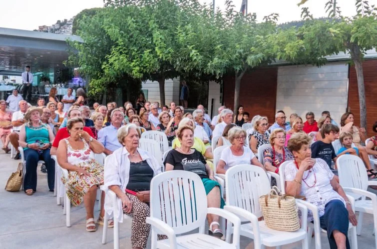 Reunión al aire libre con muchas personas sentadas en sillas, árboles y un edificio moderno en el fondo.
