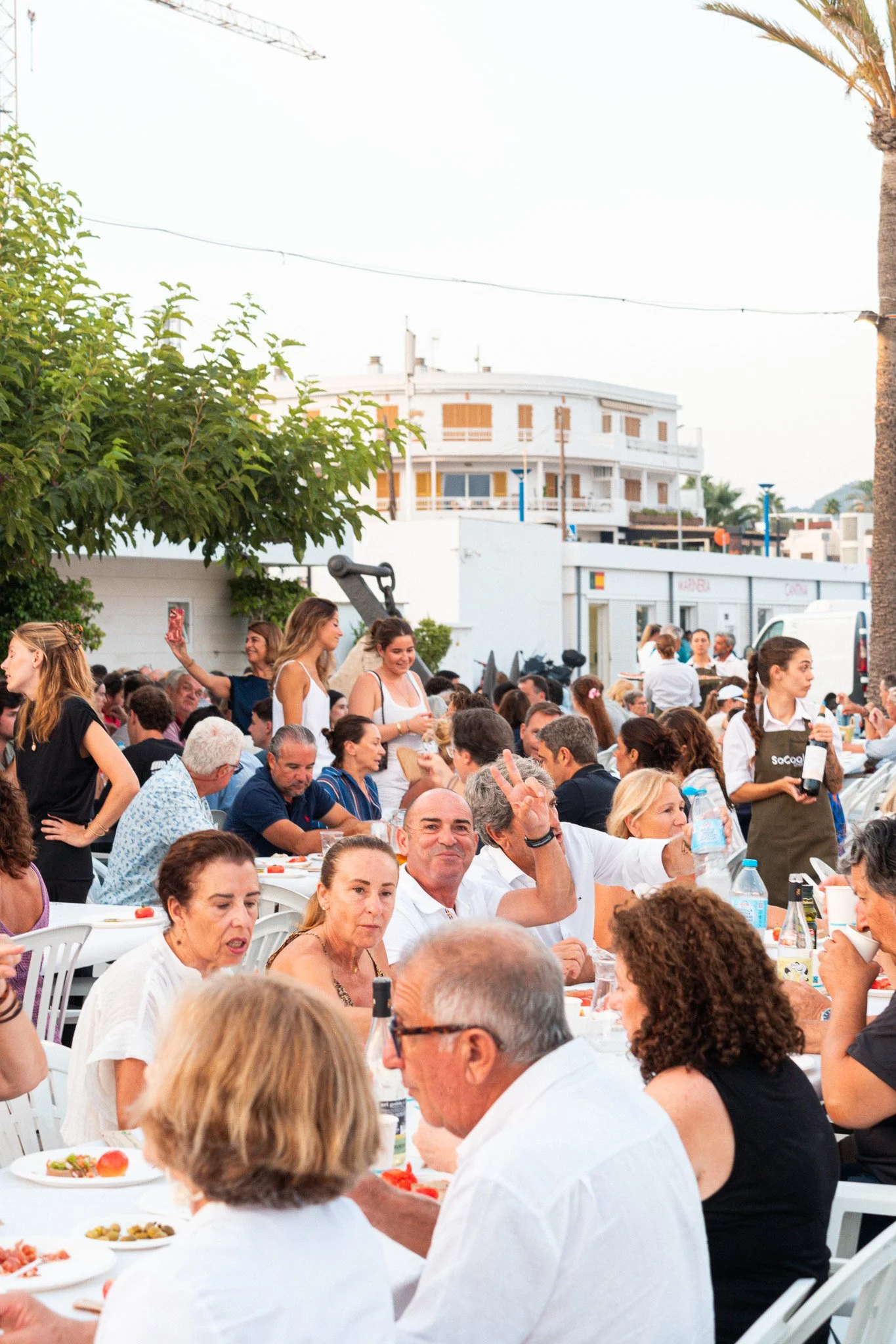Personas sentadas en una mesa al aire libre durante una comida, con comida en platos, en un ambiente festivo y social, con edificios y palmeras de fondo.