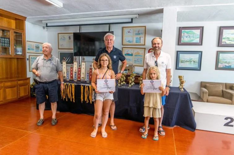 Cuatro adultos y dos niñas posando en una ceremonia de premios en una sala con cuadros en las paredes y trofeos sobre una mesa.