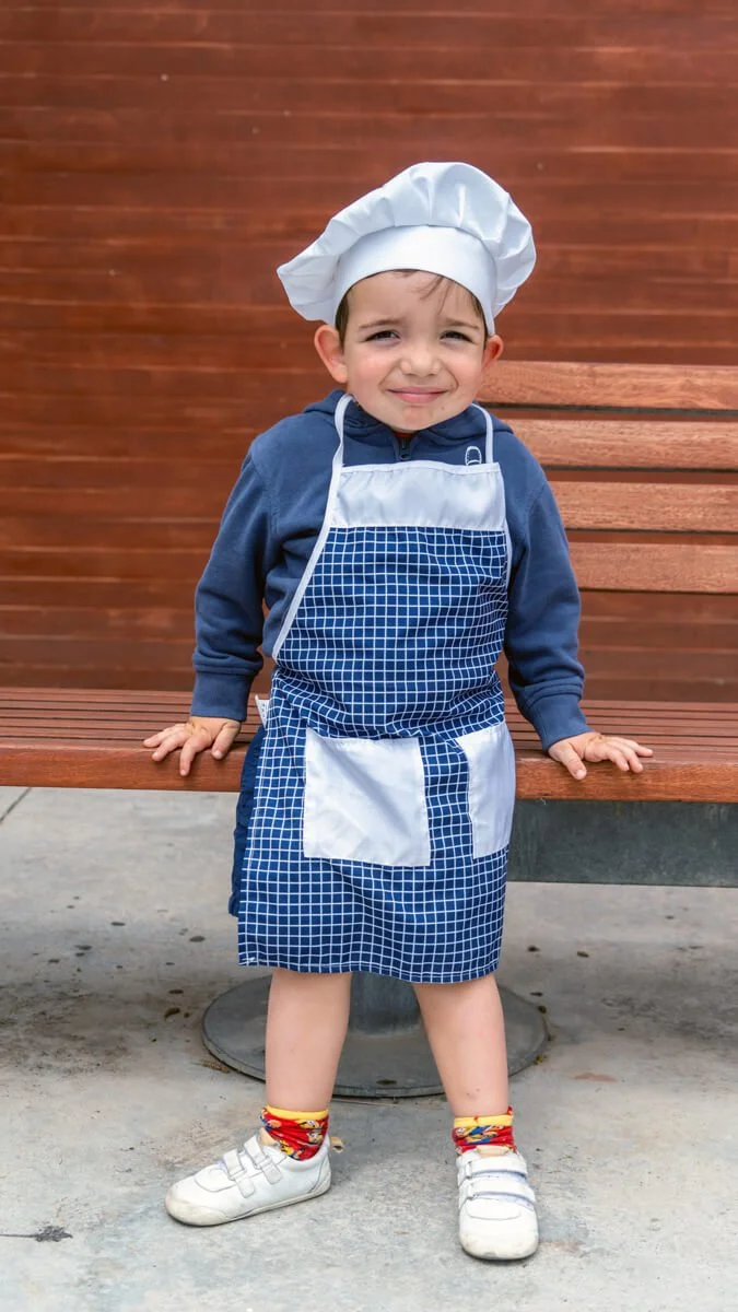 Niño pequeño con delantal de chef y gorro de chef posando en un banco de madera.