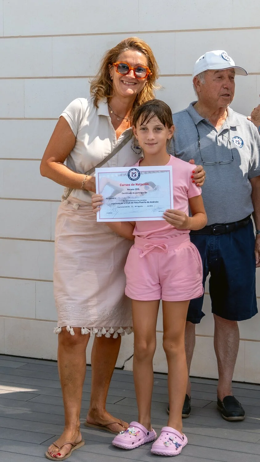 Niña sosteniendo un certificado de participación en un curso de natación, acompañada de dos adultos, en una ceremonia al aire libre.