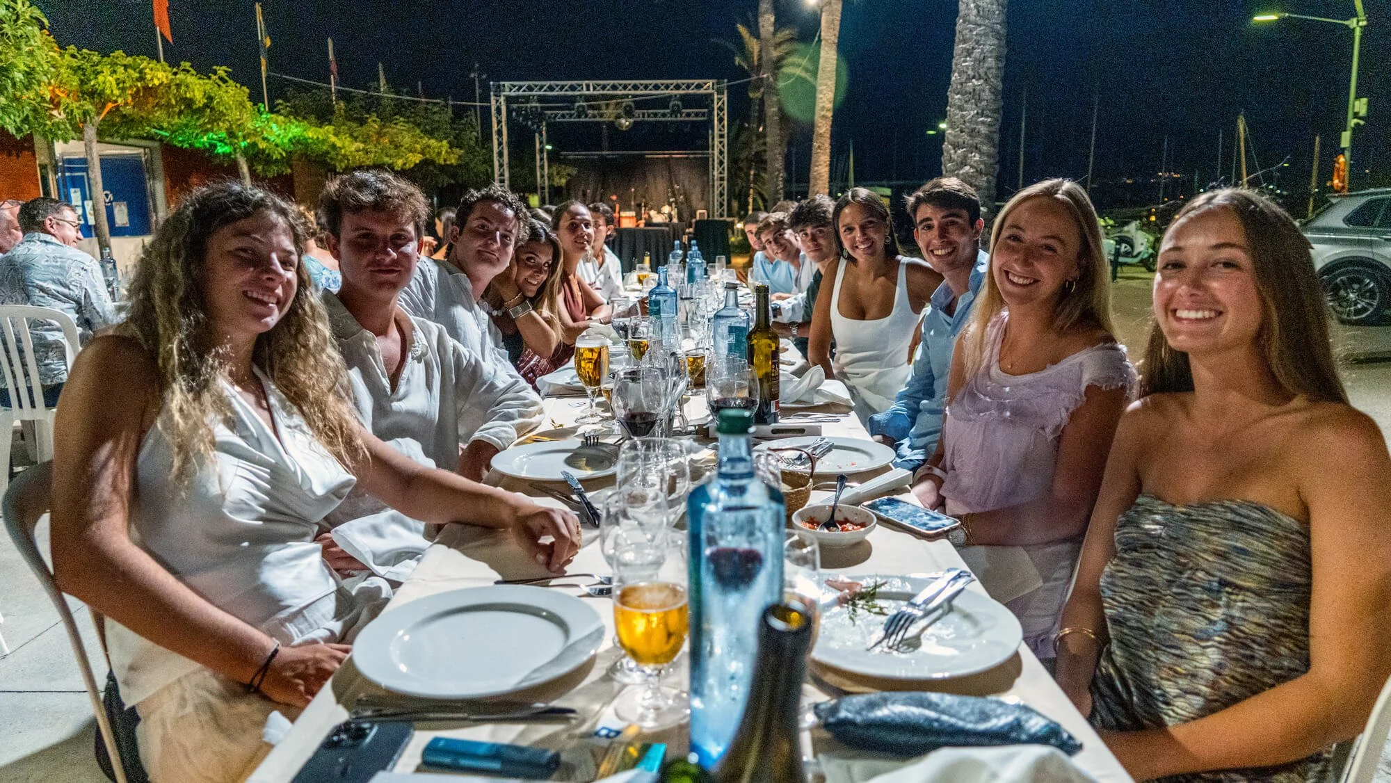 Un grupo de personas sonrientes sentadas en una mesa larga en una cena al aire libre por la noche, con decoración y escenario en el fondo.