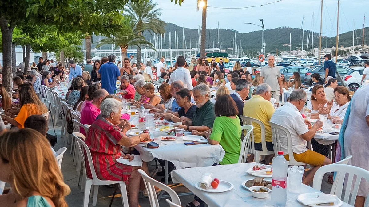 Reunión social al aire libre en un muelle con mesas llenas de comensales disfrutando una comida, con mar y yates de fondo.