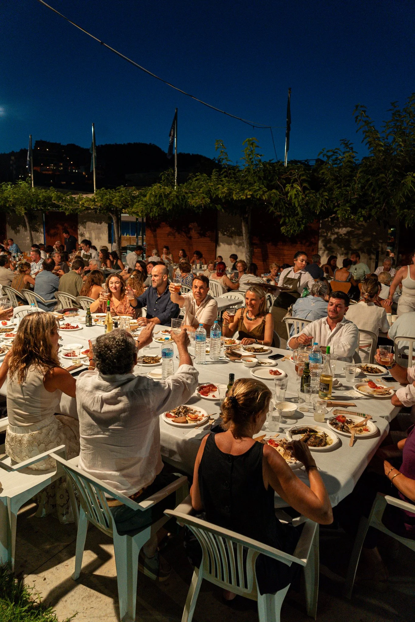 Una reunión al aire libre con muchas personas sentadas en mesas redondas, compartiendo comida y brindando en la noche, con fondo de cielo nocturno y árboles.