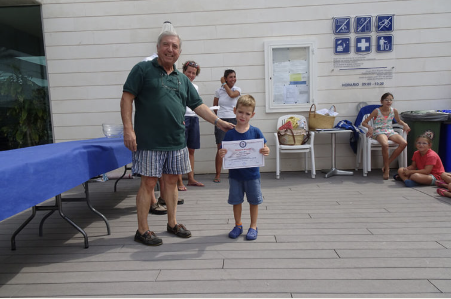 Un hombre mayor entrega un certificado a un niño en un evento al aire libre, con varias personas sentadas y de pie en el fondo y un cartel de instrucciones en la pared.