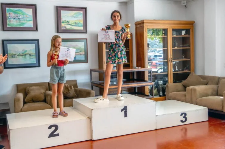 Dos niñas en un podio celebrando con trofeos y certificados en una sala con pinturas en la pared y muebles de madera