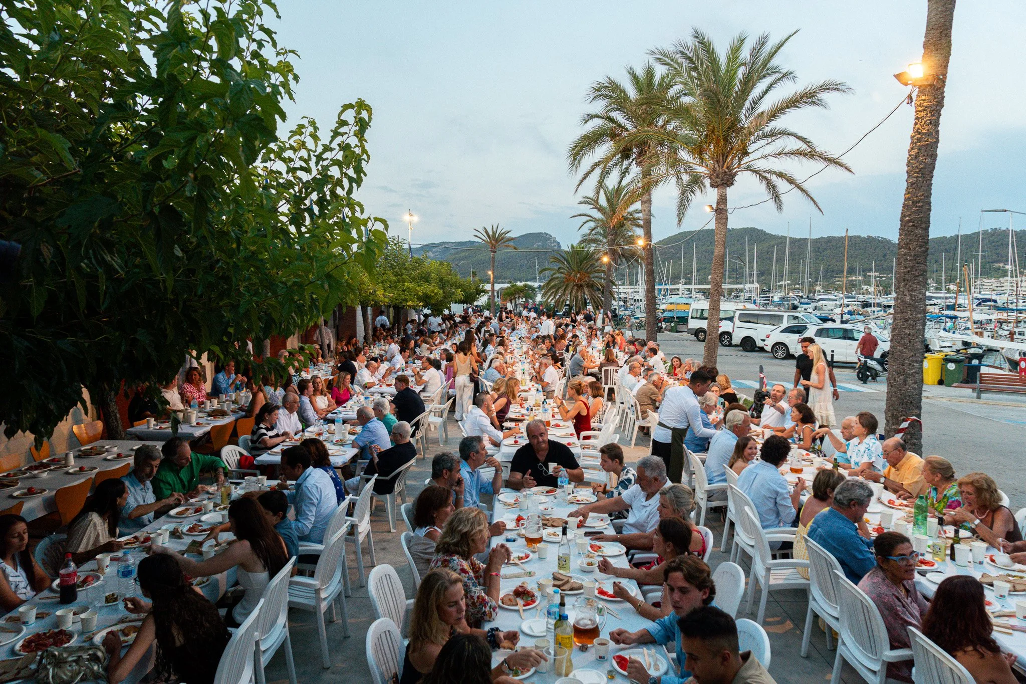 Una gran reunión en la playa con muchas mesas llenas de personas disfrutando de una comida al aire libre, con palmeras y yates en el fondo, al atardecer.