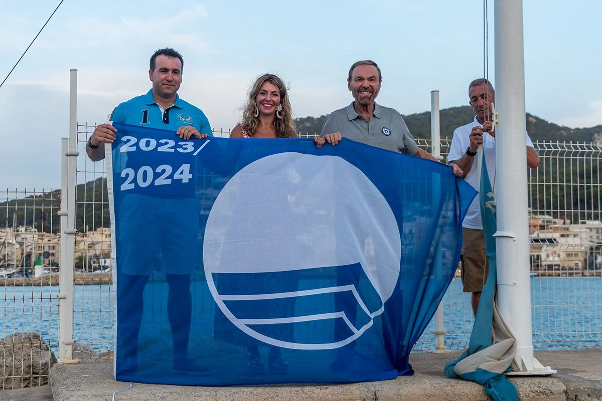 Cuatro personas sosteniendo una bandera azul con un símbolo circular y letras, en un muelle junto al agua, con montañas y una ciudad al fondo.